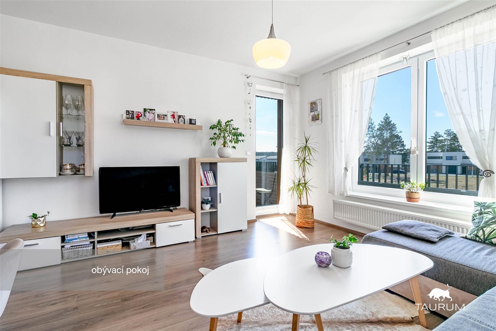 Světlý obývací pokoj s parketovou podlahou, velkými okny a moderním designem, který zajišťuje příjemnou atmosféru pro relaxaci a setkání.