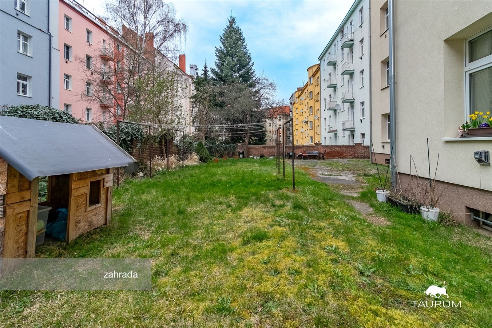 Zahrada s travnatou plochou, dřevěným zahradním domkem a okolními bytovými domy, vhodná pro relaxaci nebo děti, klidné městské prostředí.