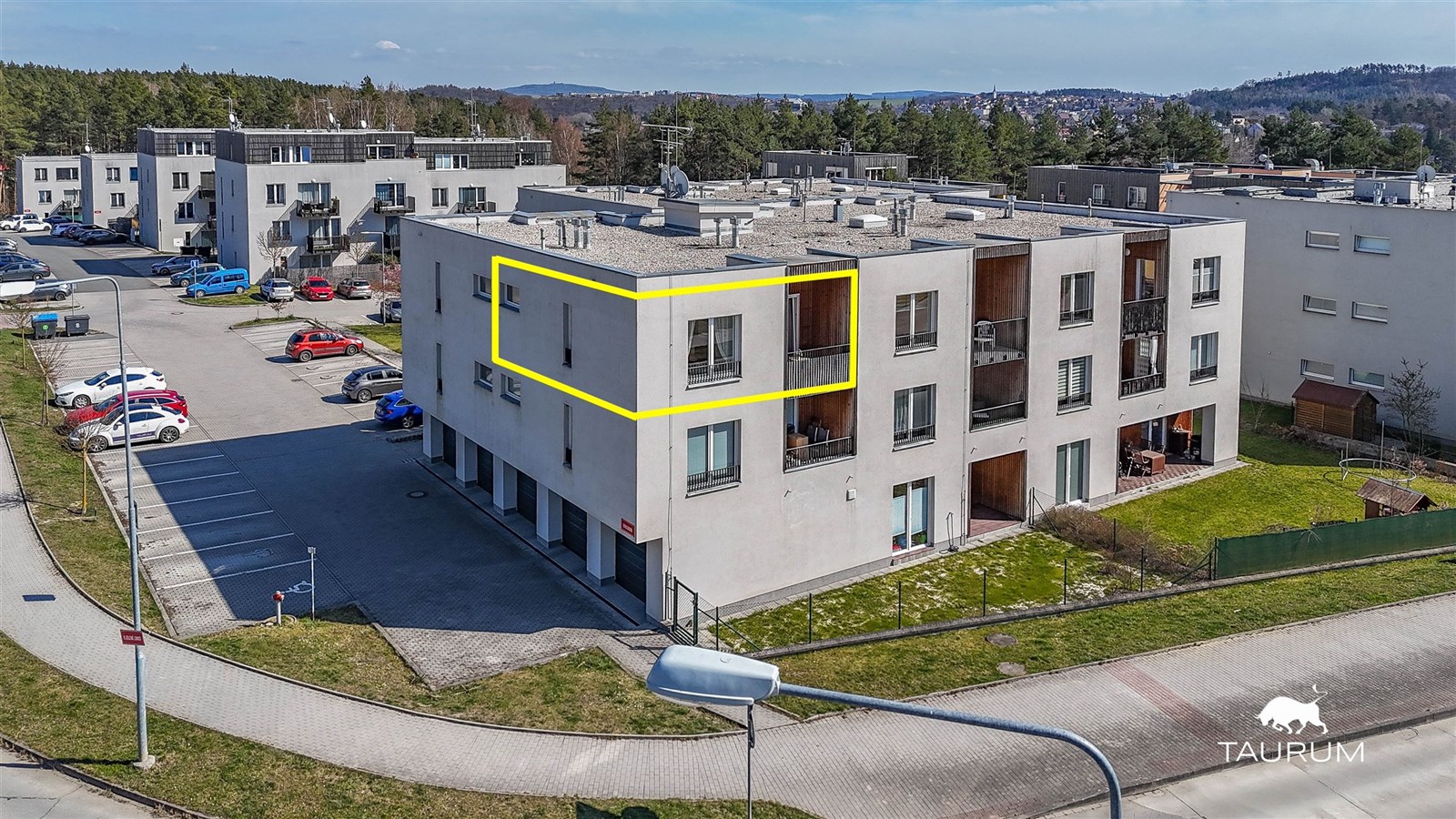 Menší bytový dům s moderním designem, světlou fasádou a plochou střechou, obklopený zelení a asfaltem.