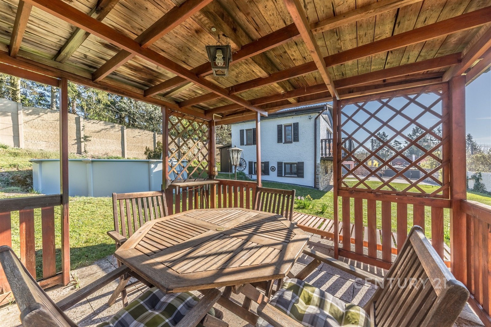 Zahradní pergola s dřevěným nábytkem a výhledem na rodinný dům. Dřevěná konstrukce, zelený trávník a nadzemní bazén vytvářejí útulné prostředí.