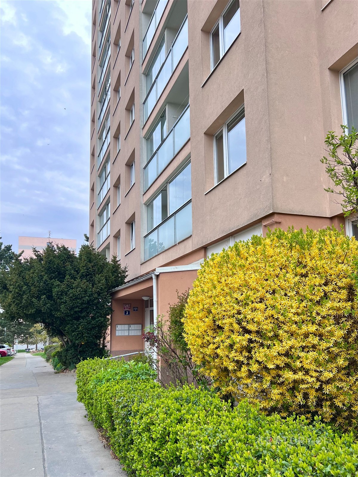 Vchod do panelového domu s upravenou zelení, moder