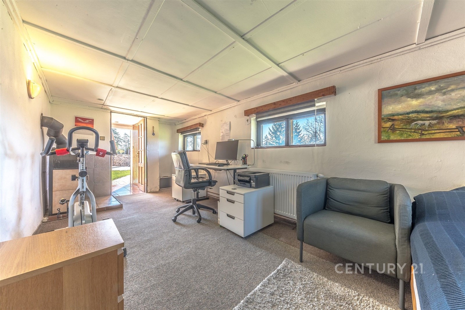 Domácí kancelář s kobercovou podlahou, světlá místnost s okny na zahradu, moderně zařízené s pracovním stolem a pohodlným křeslem.