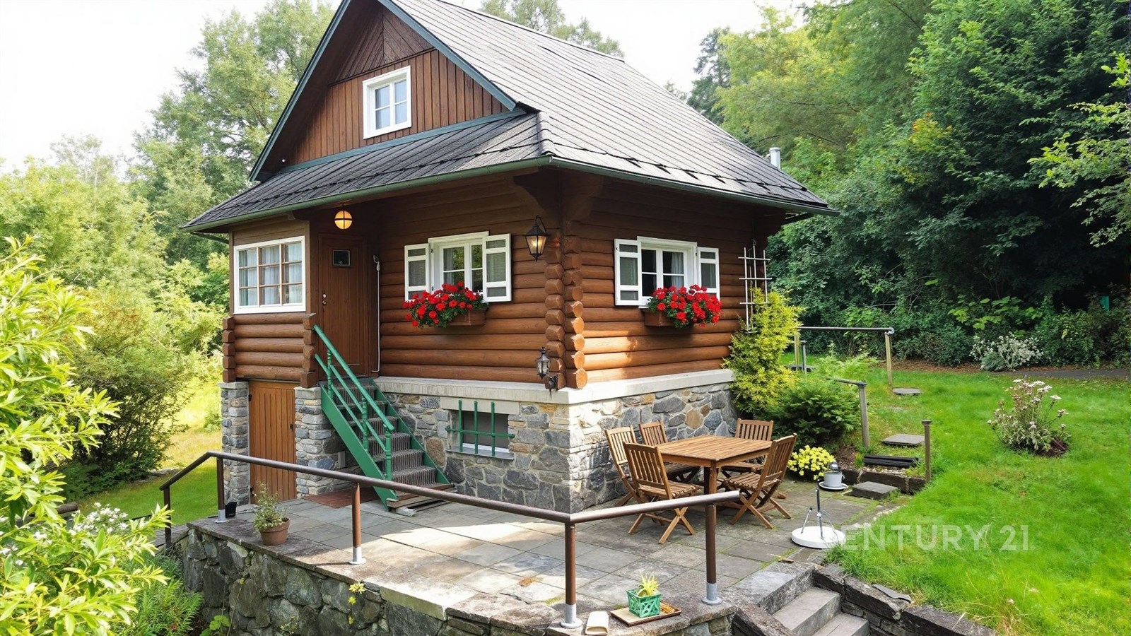 Dřevěná chata s kamennými základy, obklopená zelení, nabízí terasu s venkovním posezením. Tradiční rustikální styl doplňují květinové truhlíky.