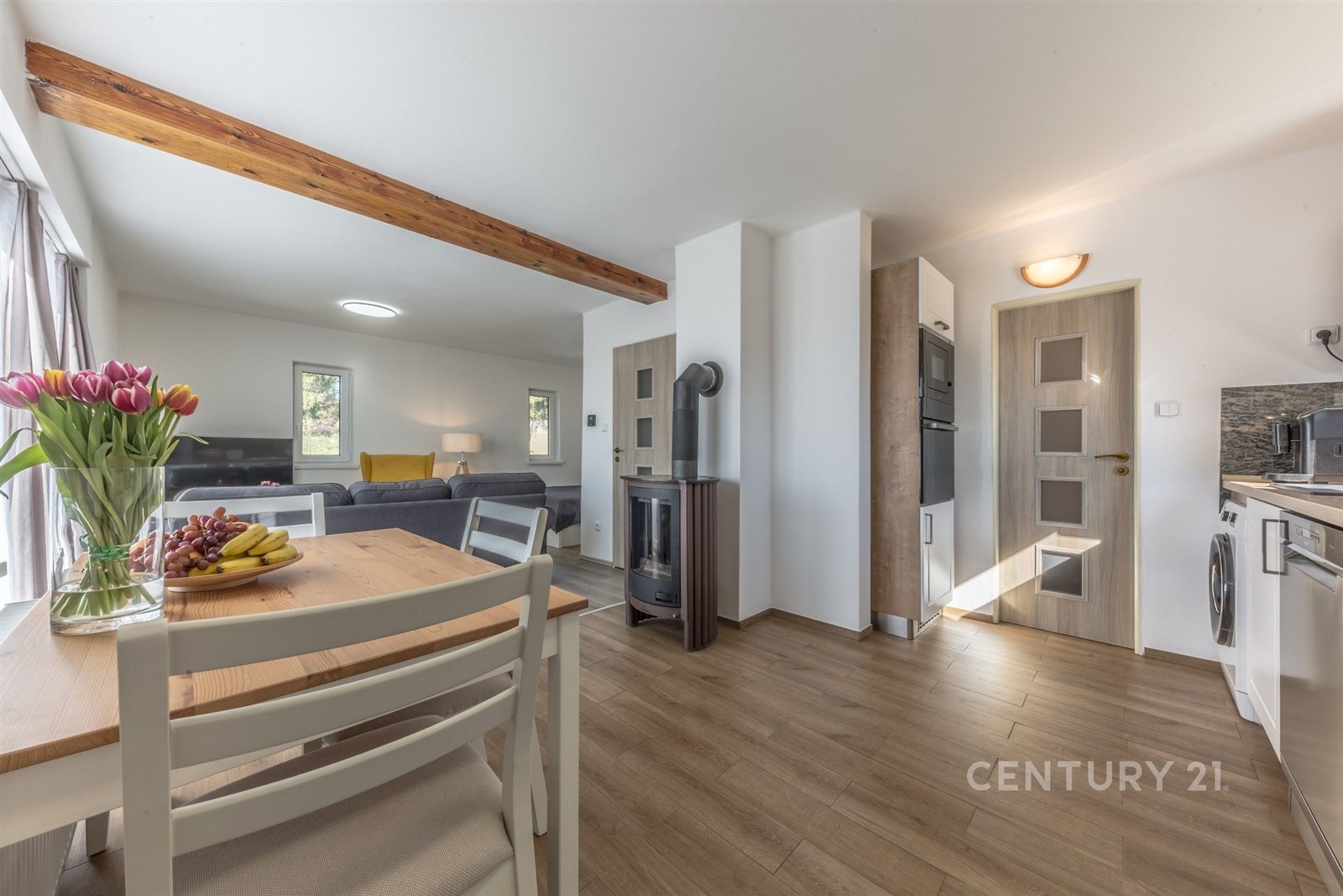 Světlá jídelna s dřevěným stolem, parketová podlaha, moderní spotřebiče, krbová kamna a velká okna dodávají prostoru moderní vzhled.