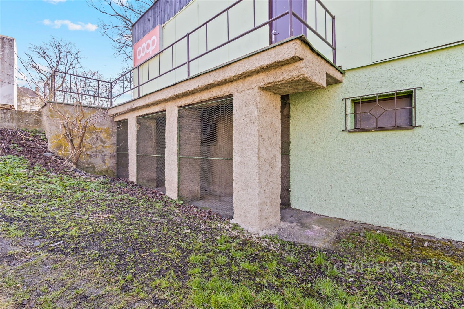 Přiléhající prostor s betonovými sloupy a hrubou omítkou, vhodný pro úpravy, vedle zatravněného pozemku, ideální pro skladování nebo dílnu.