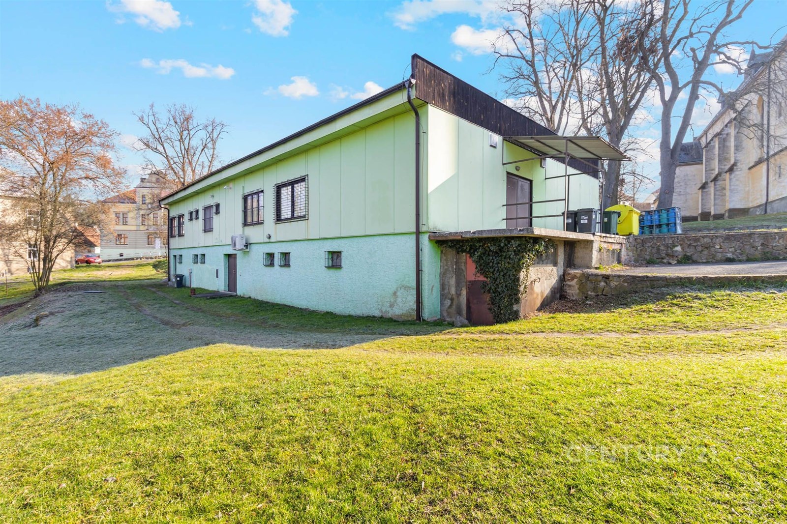 Světle zelená budova s moderními okny a plochou střechou na prostorném pozemku s trávníkem, ideální pro komerční využití nebo přestavbu.