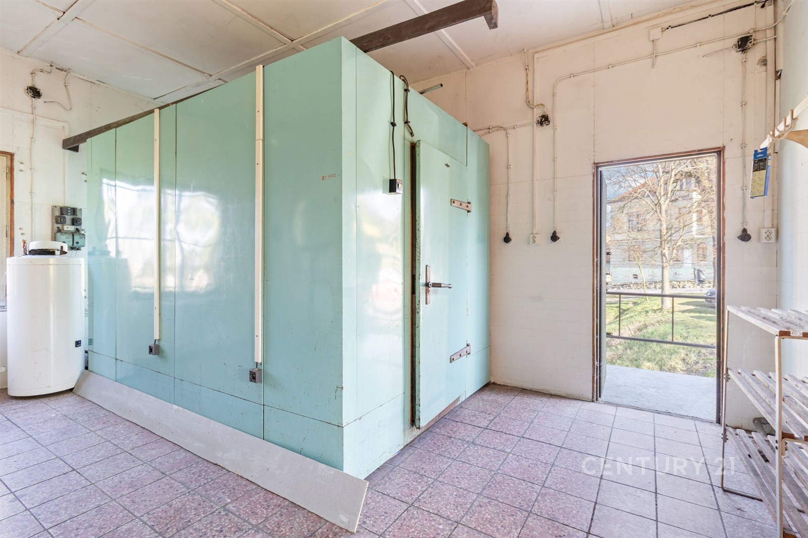 Průmyslový prostor s dlažbou, světlý interiér, velká chladicí jednotka, přístup na venkovní plochu, vhodné pro komerční využití.