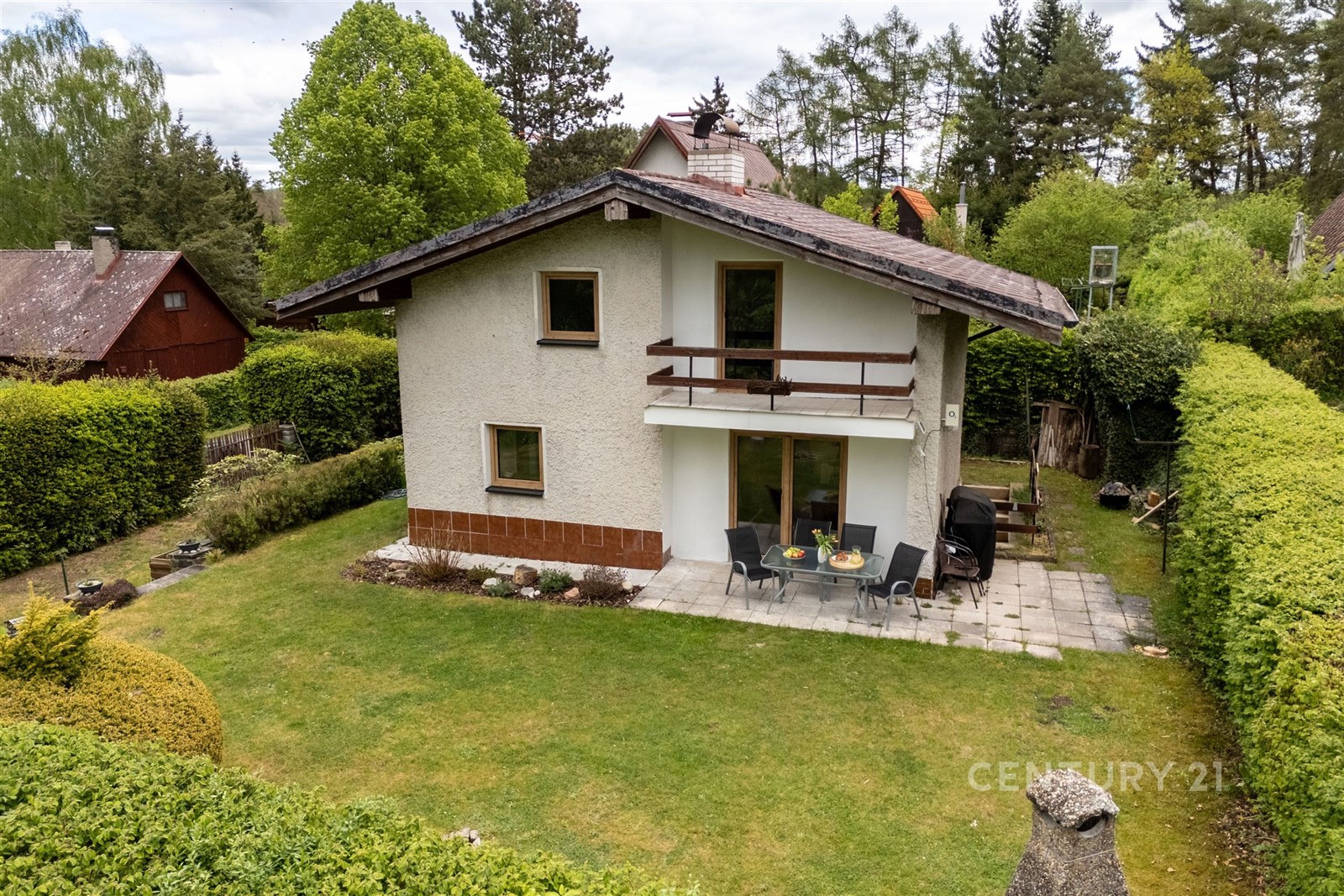 Rodinný dům s terasou v zahradě, obklopený zelení. Dům má dřevěný balkon a je v klidné lokalitě, ideální pro relaxaci.