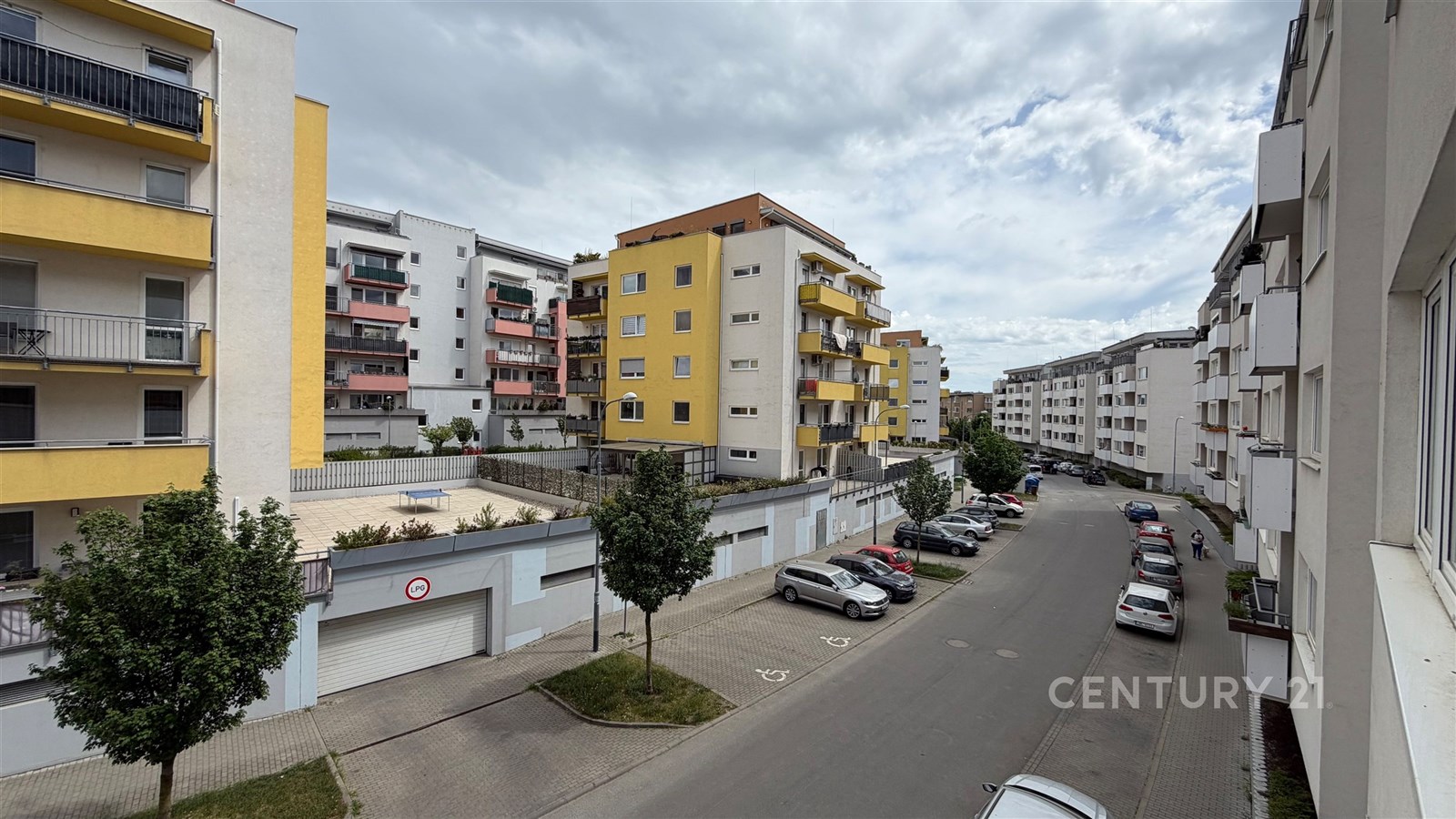 Pohled z okna na moderní sídliště s barevnými fasádami, balkonky a zaparkovanými auty.