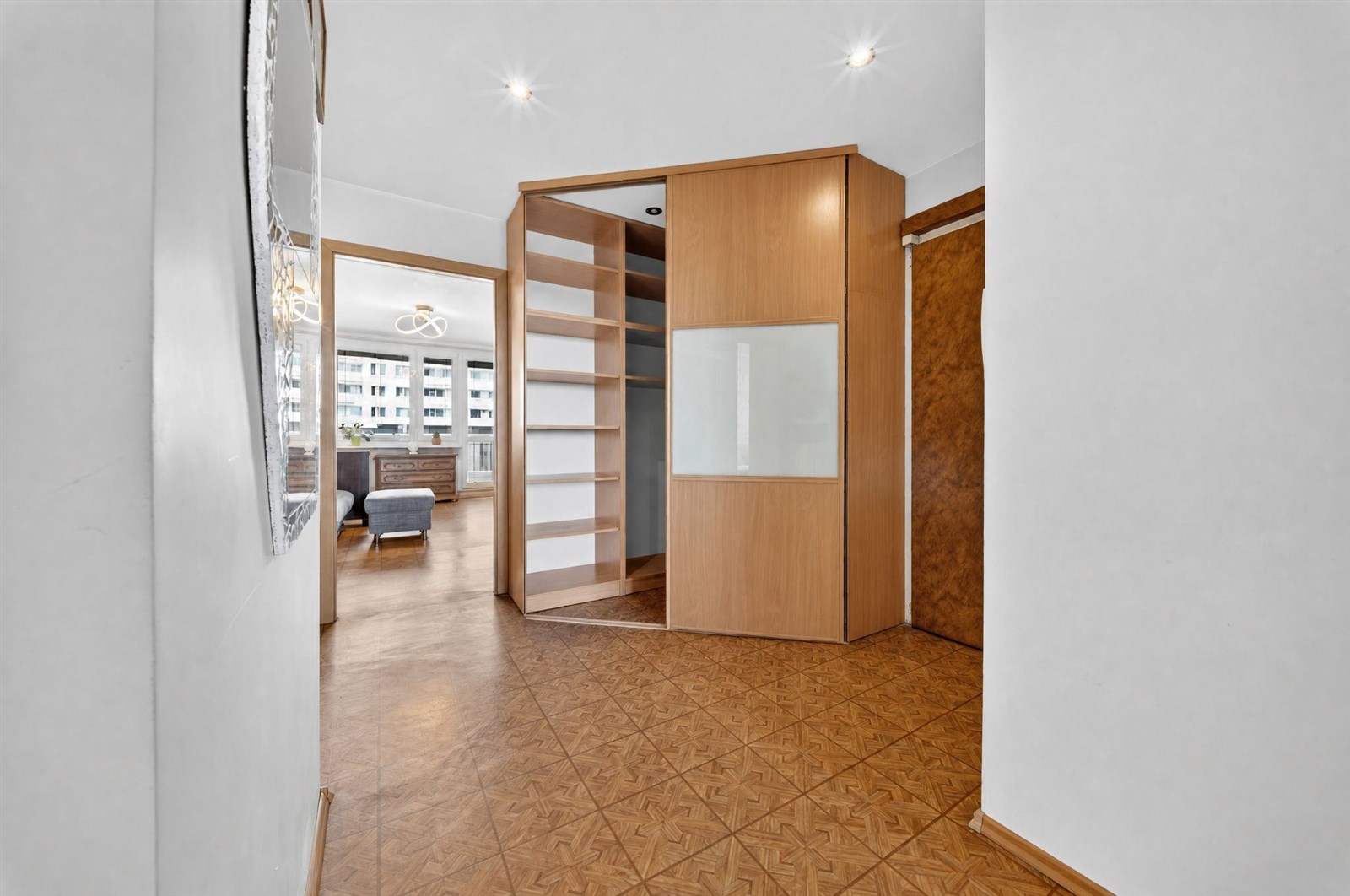 Chodba s parketovou podlahou a vestavěnými skříněmi, moderní design; velká okna poskytují přirozené světlo do sousedního prostoru.