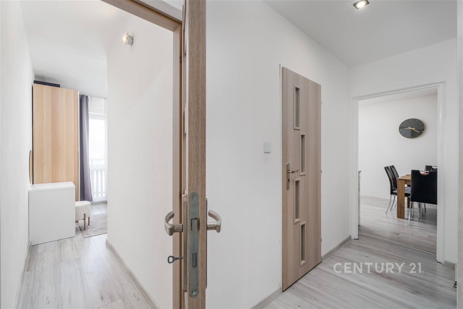 Světlá chodba s laminátovou podlahou a moderními dveřmi, vedoucí do jídelny s minimalistickým nábytkem a velkými okny pro přirozené světlo.