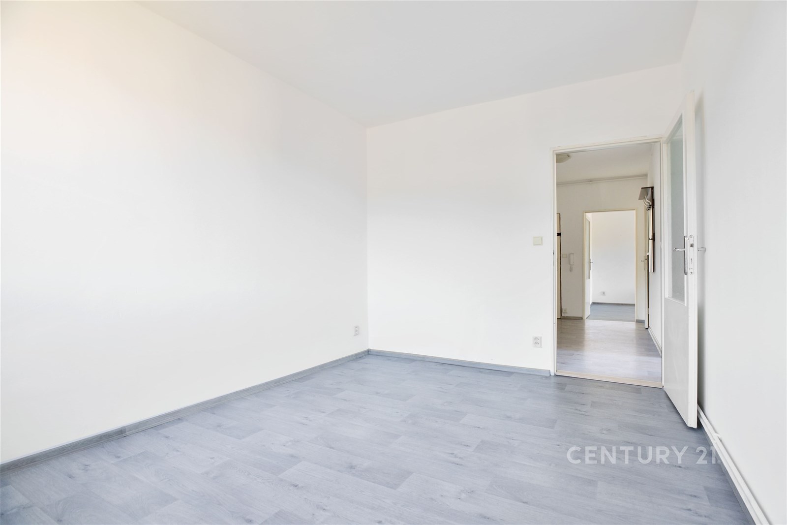 Světlý pokoj s moderní parketovou podlahou, bílé stěny a dveře vedoucí do chodby, ideální pro snadnou personalizaci interiéru.