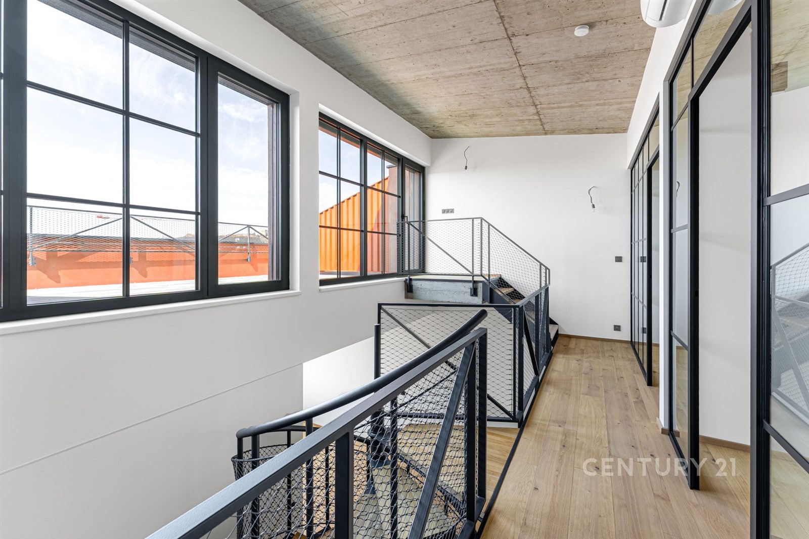 Světlá chodba s velkými okny, moderním průmyslovým stylem a dřevěnou podlahou. Skleněné stěny s kovovými rámy dodávají industriální vzhled.