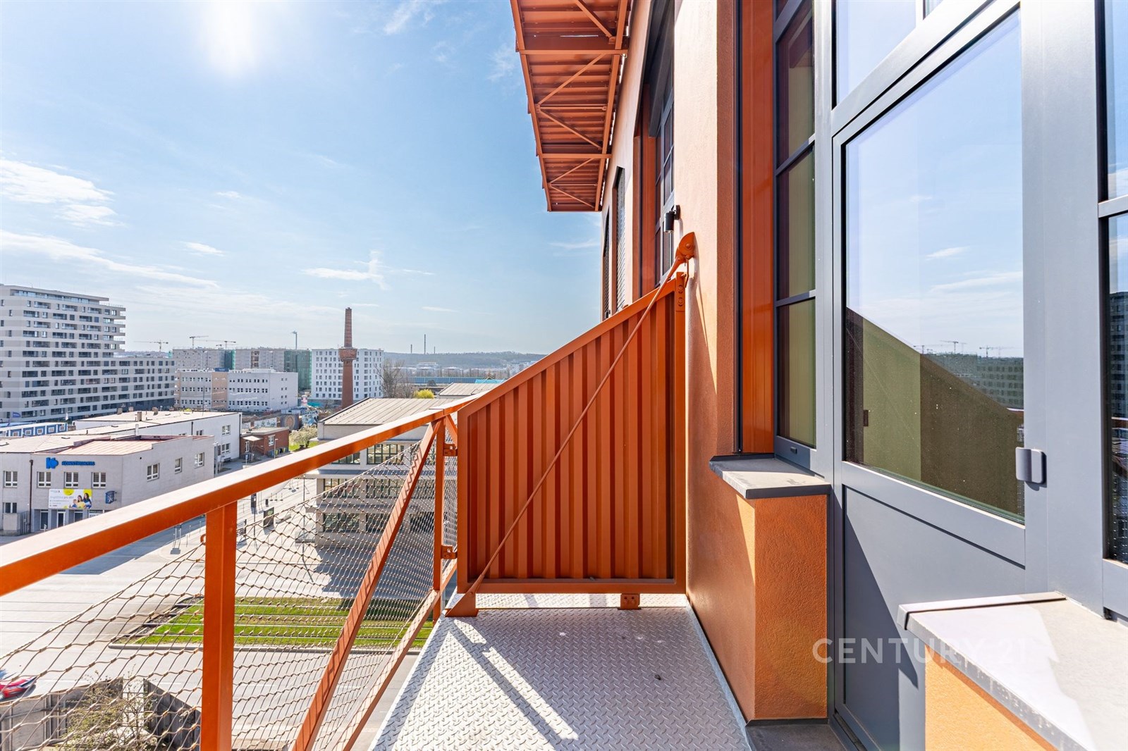 Balkon s moderními kovovými zábradlími, výhledem na město a vstupem přes prosklené dveře, ideální pro slunné dny.