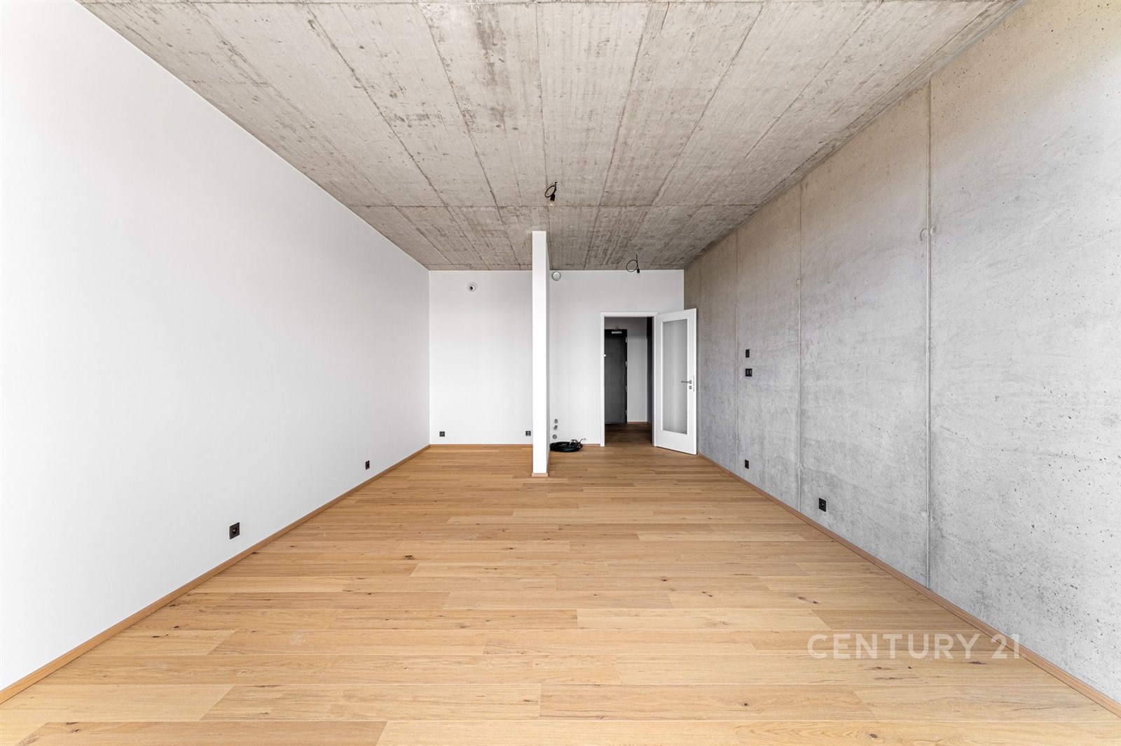 Prázdný obývací pokoj s betonovým stropem, parketovou podlahou a bílými stěnami, nabízí moderní styl a připravenost k osobnímu uzpůsobení.