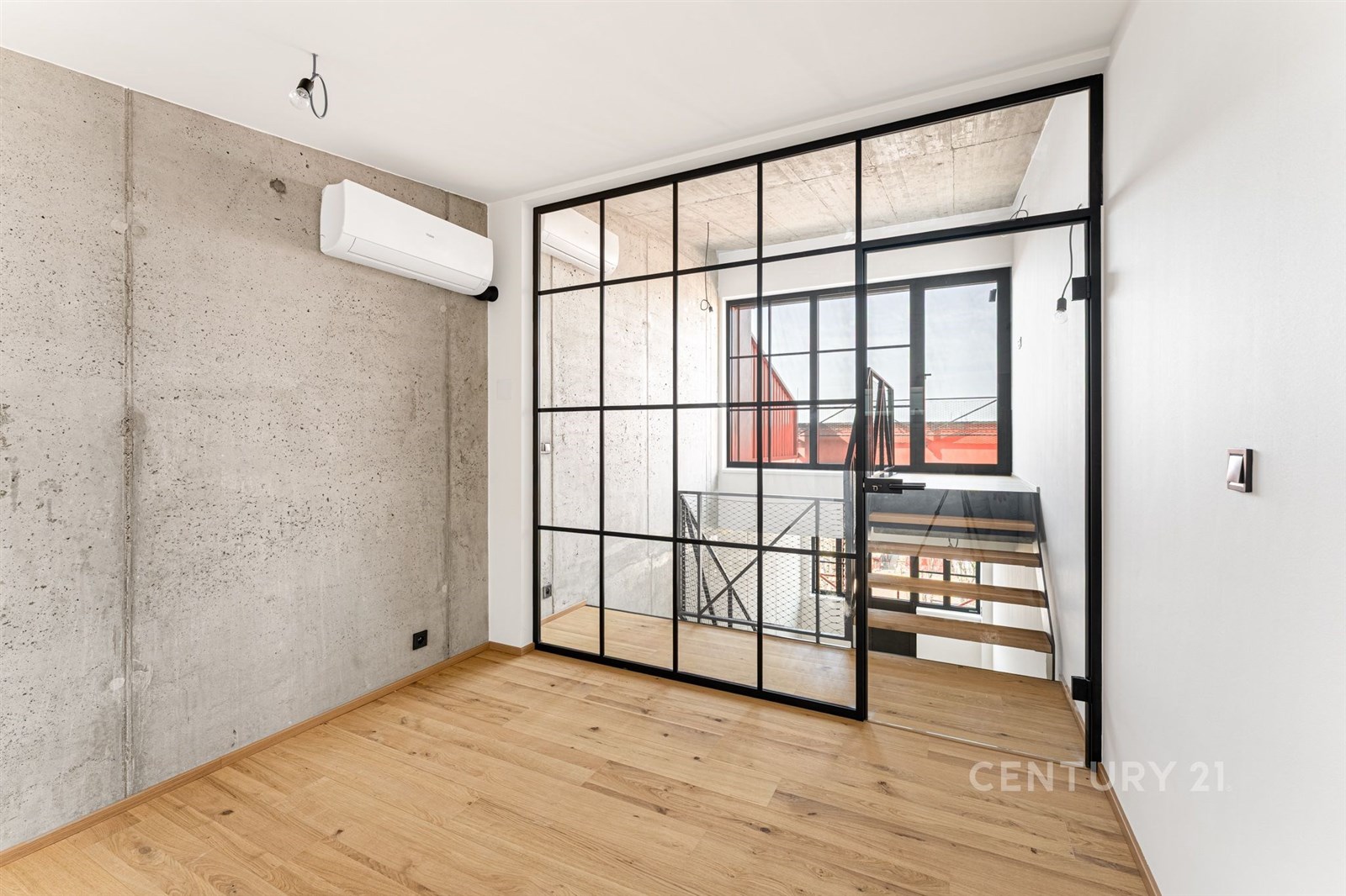 Průmyslově stylizovaná místnost s parketovou podlahou a betonovou stěnou, oddělena prosklenými dveřmi, poskytuje moderní a světelný interiér.