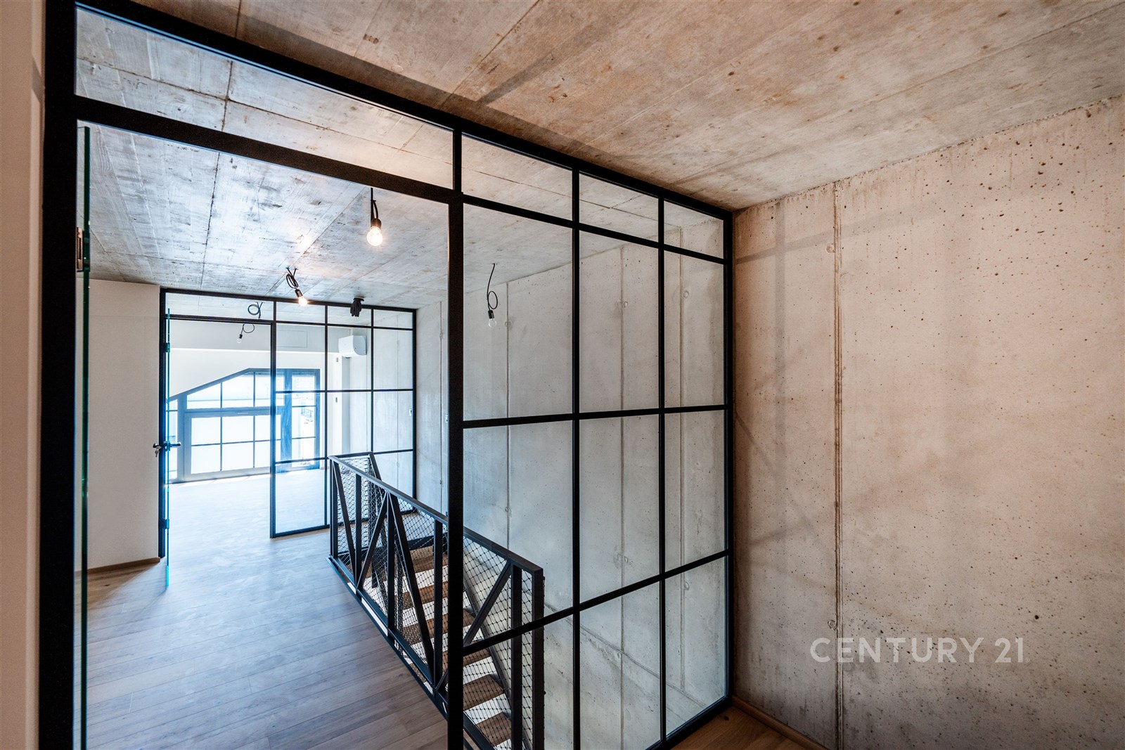 Moderní chodba s průmyslovým designem, betonové stropy, skleněné příčky a dřevěná podlaha, propojující otevřené prostory s přirozeným světlem.