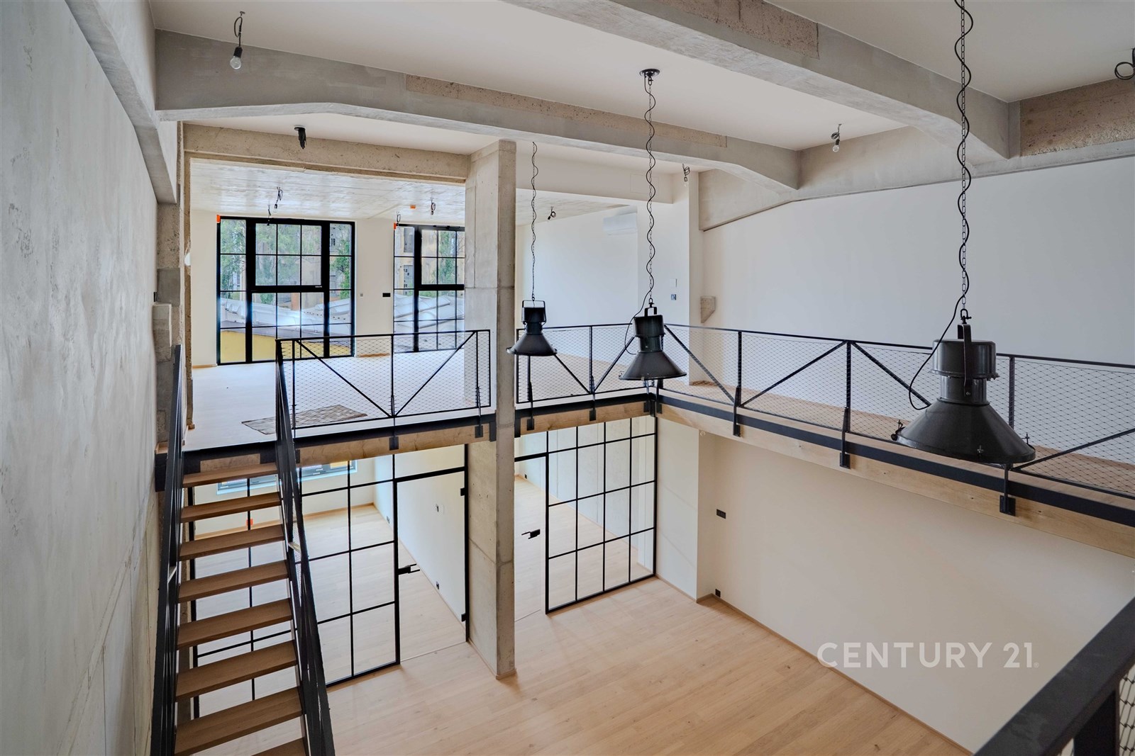 Moderní otevřený prostor s parketovou podlahou, velkými okny a industriálními prvky. Světlý interiér s kovovým zábradlím a schodištěm.