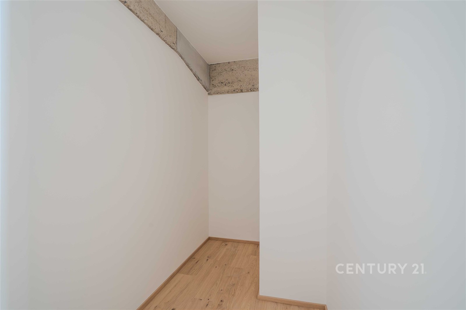 Malá místnost s parketovou podlahou, bílými stěnami a odkrytým betonovým trámem, ideální pro úložný prostor nebo komoru.