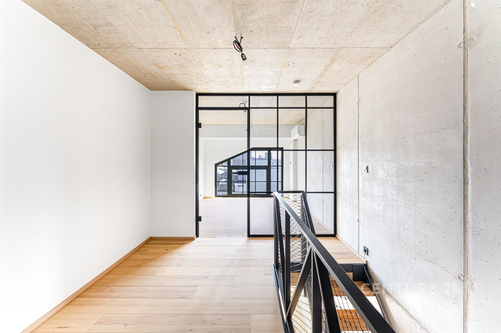 Moderní hala s parketovou podlahou, betonovým stropem a skleněnými dveřmi. Světlý interiér nabízí hlavní vstup do dalších prostor.