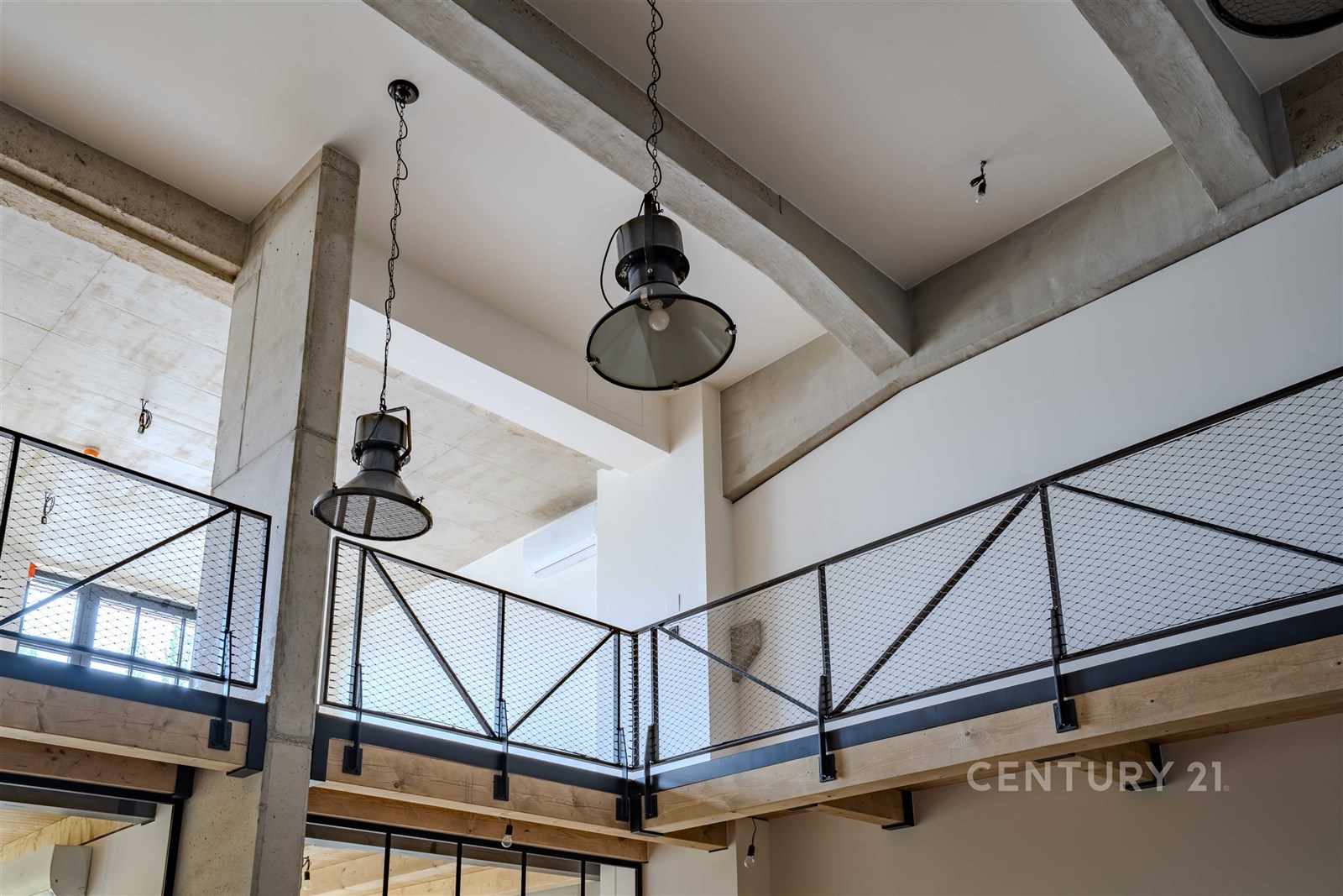 Moderní loftový prostor s vysokými stropy, betonovými prvky a kovovým zábradlím. Průmyslový styl a velká okna poskytují dostatek světla.