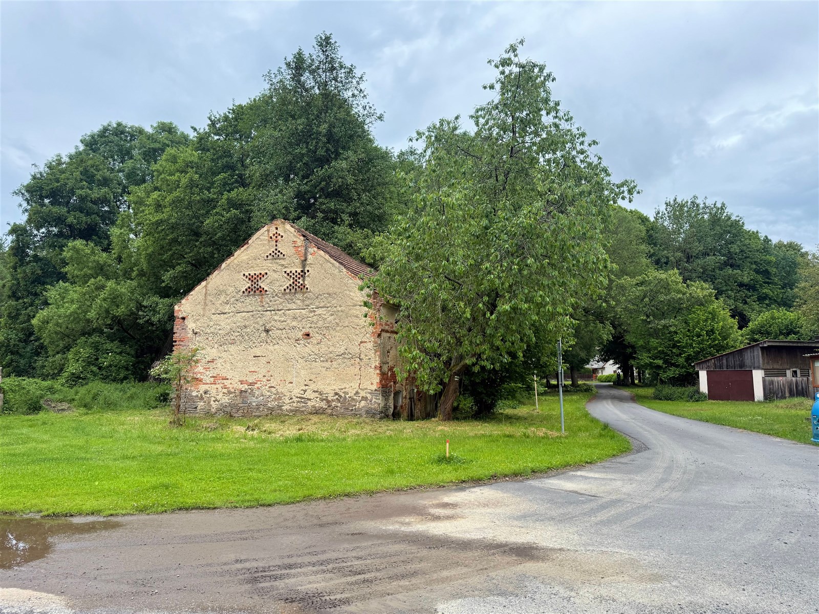 Prázdný pozemek s částečně zchátralou budovou, obklopený zelení a stromy, vhodný pro rekonstrukci nebo nový projekt, v klidné lokalitě.