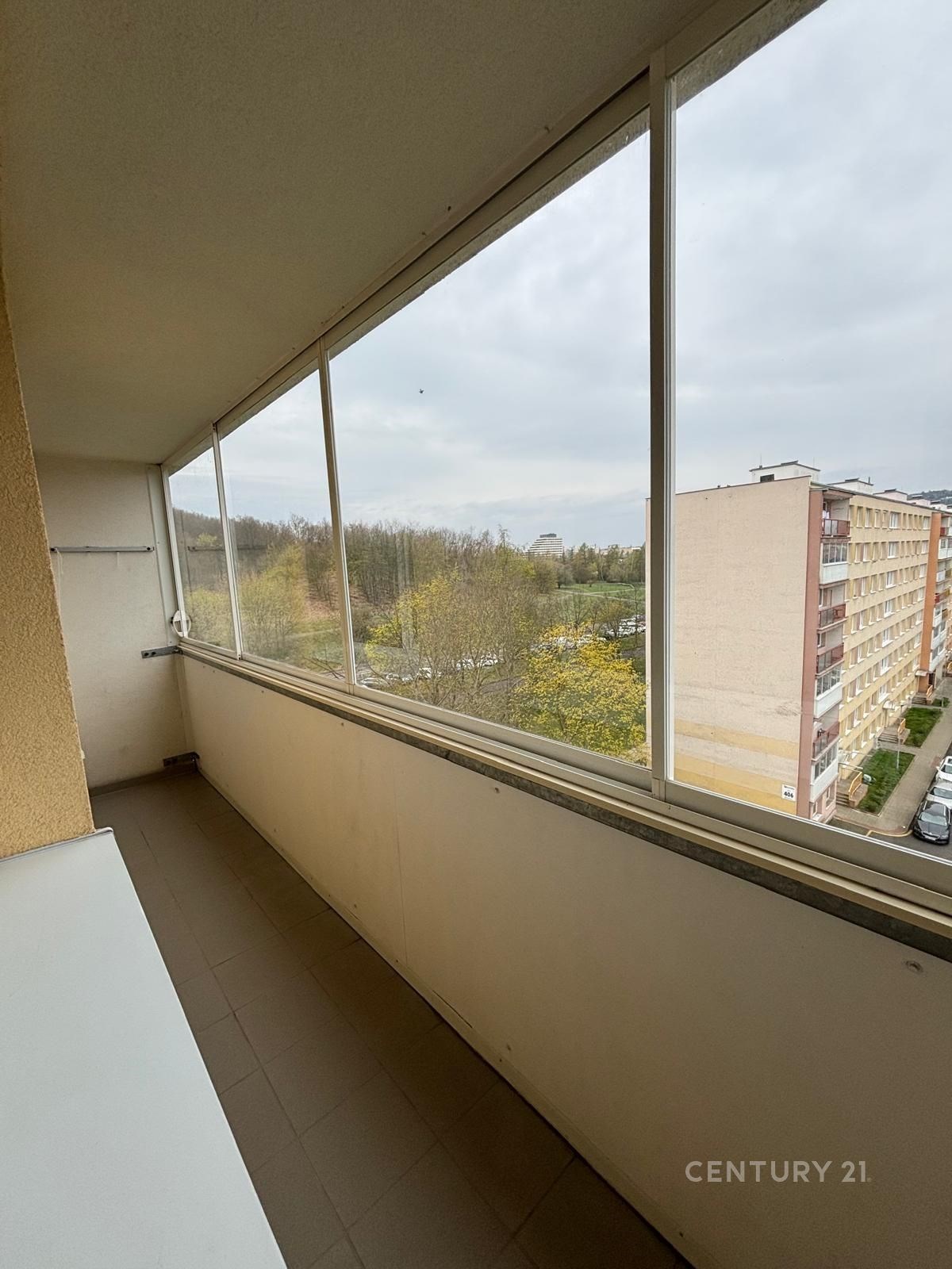 Zasklená lodžie s dlažbou, výhledem na zeleň a okolní panelové domy, vhodná pro relaxaci nebo pěstování rostlin.