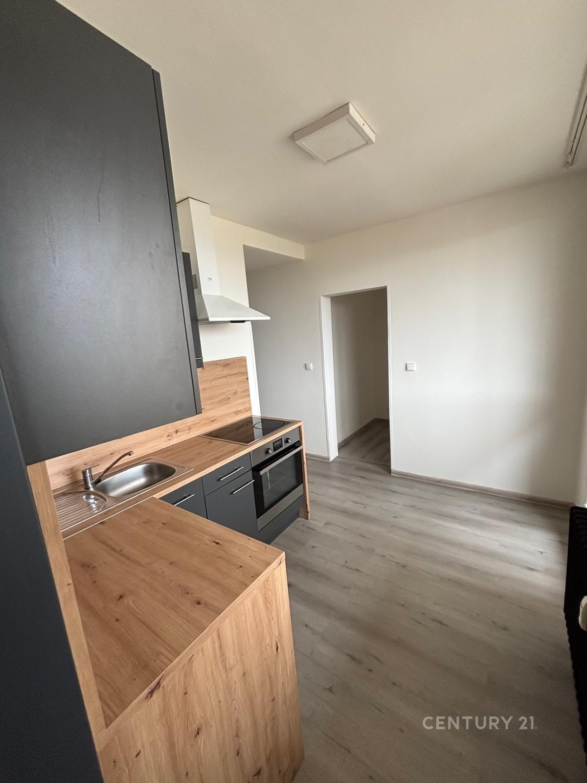 Kompaktní kuchyň s tmavou linkou, dřevěným obložením a moderními spotřebiči, světlá podlaha, minimalistický styl.