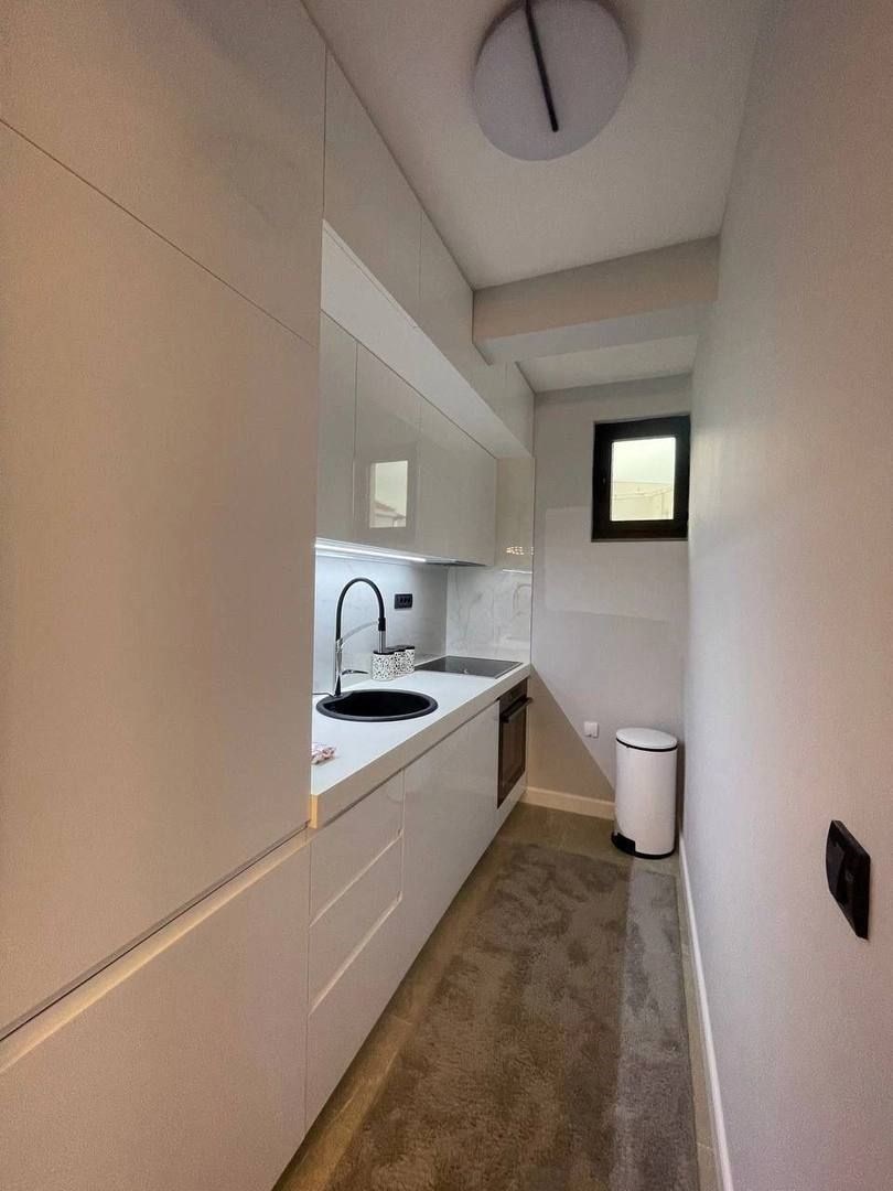 Úzká kuchyň s moderním designem, bílými skříňkami a vestavěnými spotřebiči, dlažba na podlaze, světlá s malým oknem pro přirozené světlo.