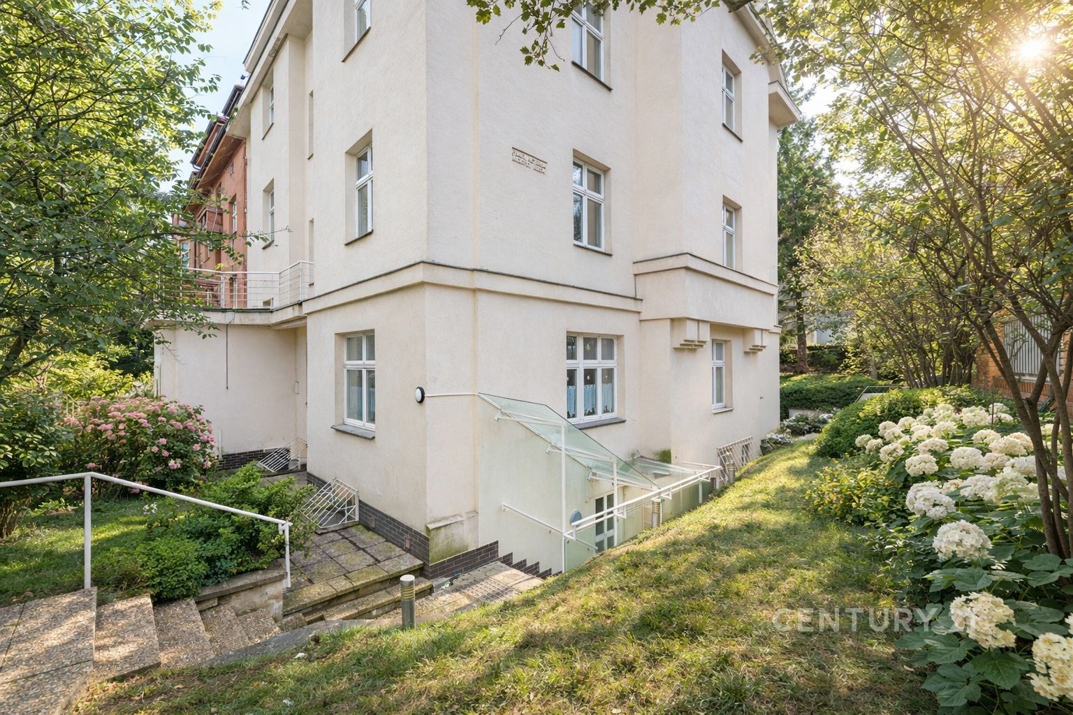 Rohová budova s bílou fasádou, obklopená upravenou zahradou s okrasnými keři a květinami, nabízí klidné okolí a dobrou dostupnost.