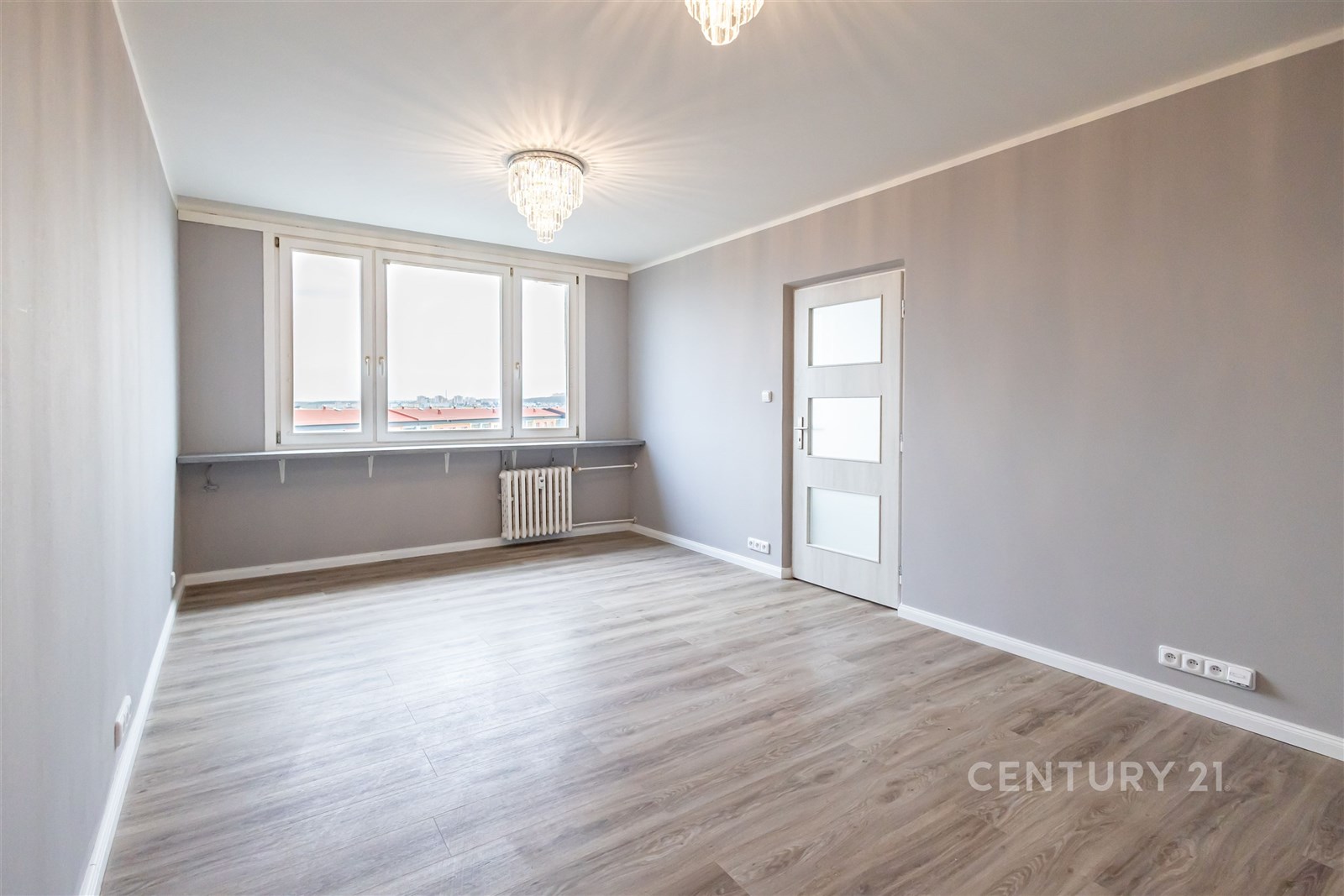 Světlý obývací pokoj, moderní parketová podlaha, velká okna, zrekonstruovaný interiér, elegantní světla, šedé stěny, čistý prostor.