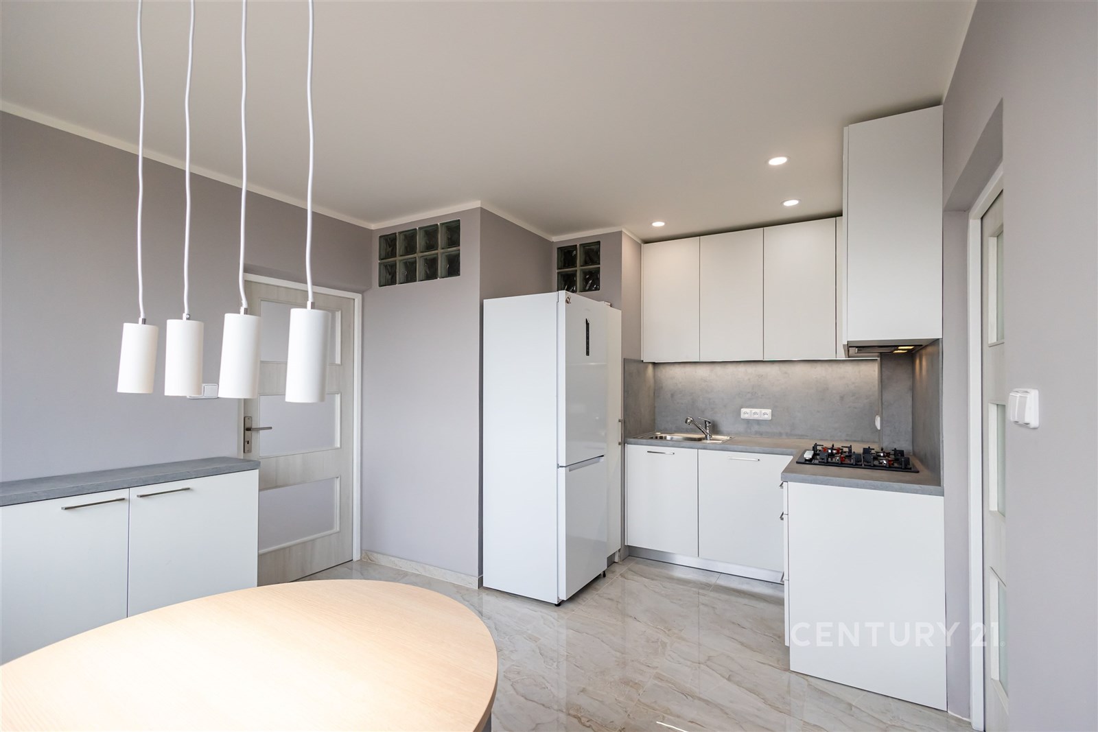 Moderní kuchyň s bílým nábytkem, mramorovou dlažbou a vestavěnými spotřebiči, světlá díky velkým oknům a minimalistickému designu.