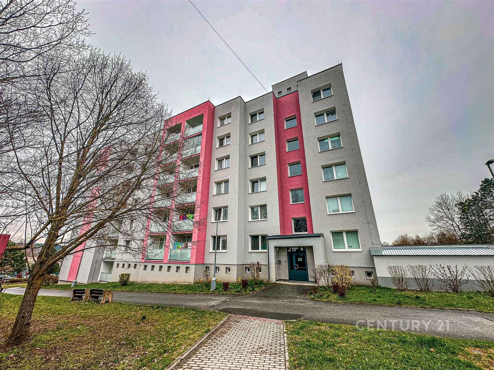 Exteriér panelového bytového domu, šedá a růžová fasáda, šest podlaží, udržovaná zeleň, klidná lokalita, modernizovaný vzhled