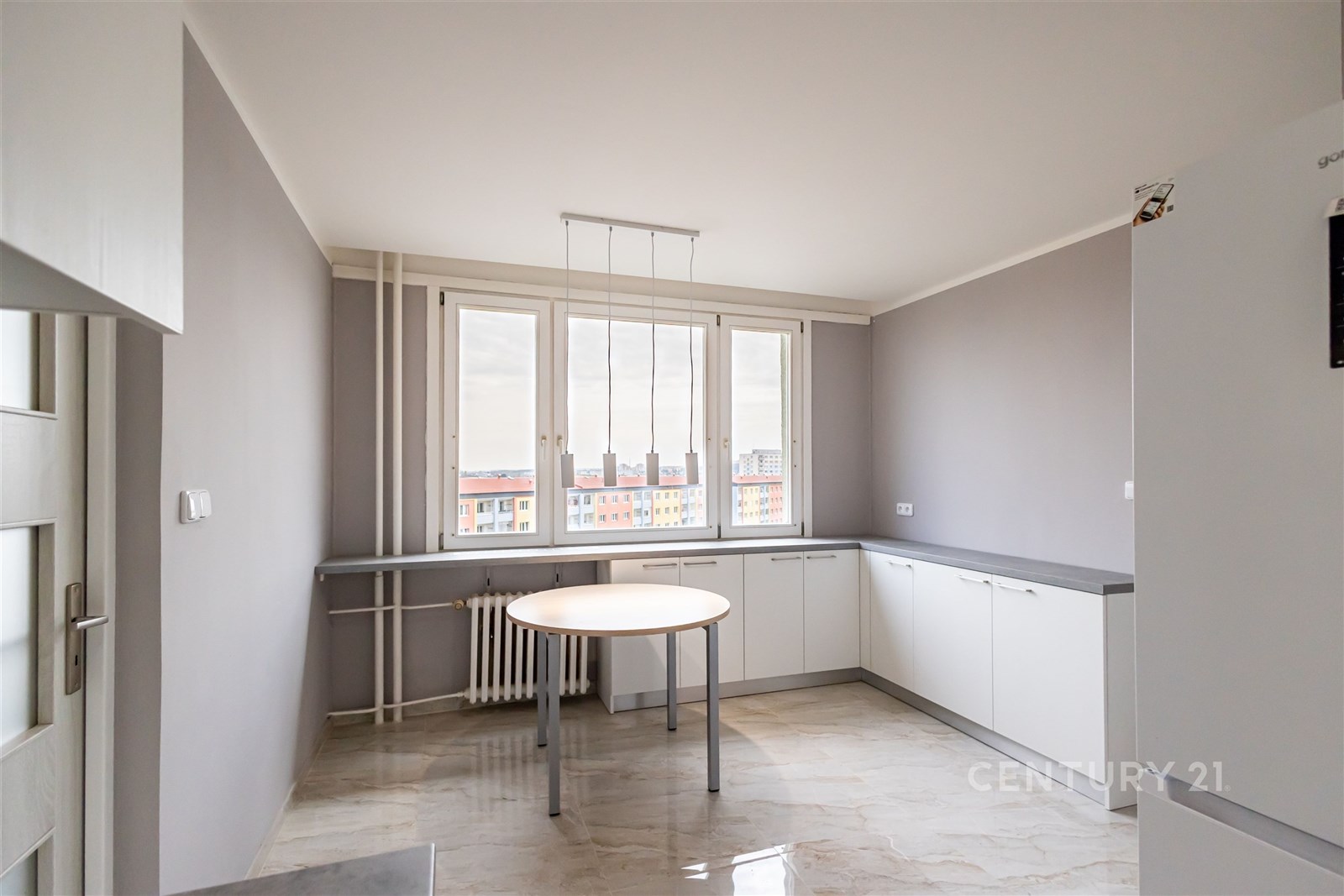 Moderní kuchyň s velkými okny, světlými skříňkami a dlažbou, nabízí prostorné uspořádání a dostatek denního světla.