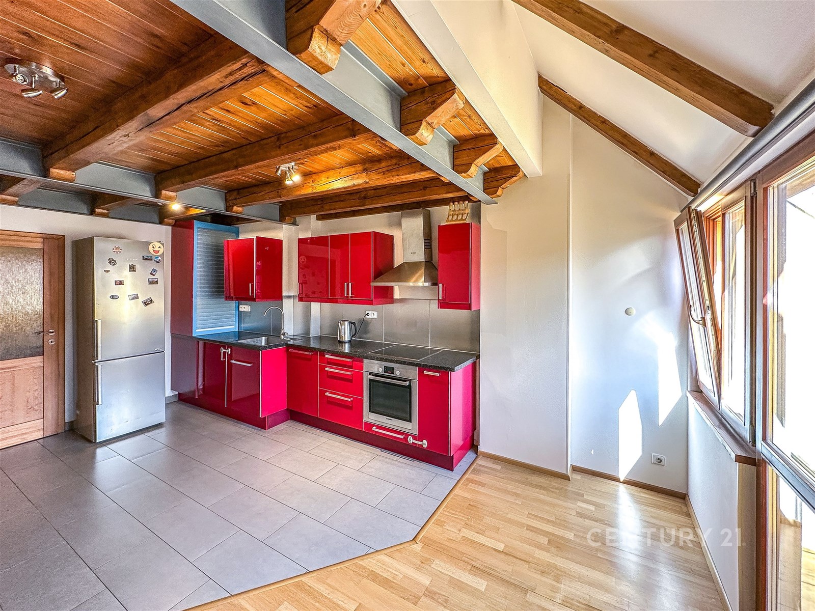 Moderní kuchyň s červenými lesklými skříňkami, nerezovými spotřebiči a dlažbou, propojená se světlým obývacím prostorem s parketovou podlahou.