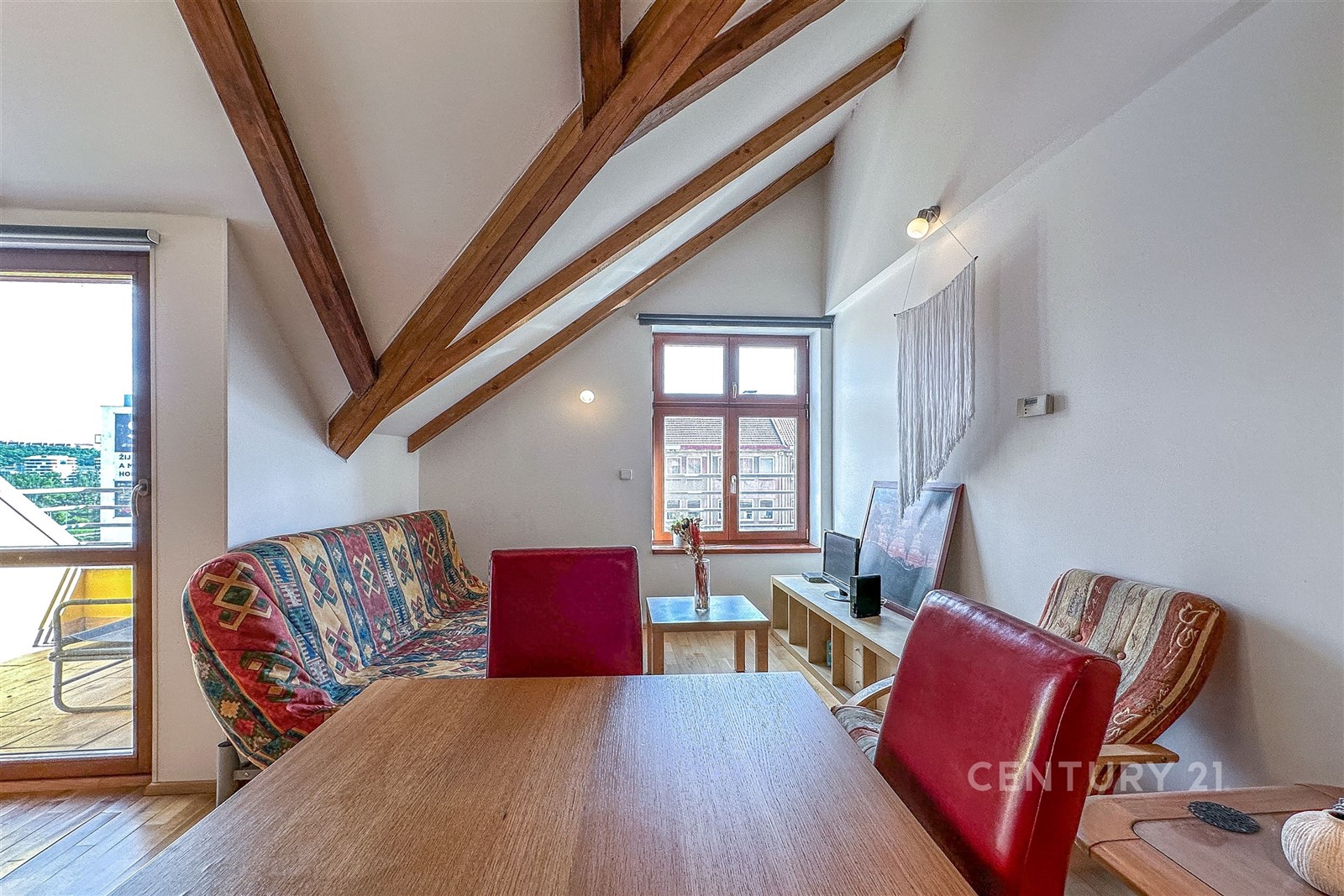 Podkrovní obývací pokoj s dřevěnými trámy, velká okna a stylový interiér, který zahrnuje světlý dřevěný nábytek a barevné sezení.