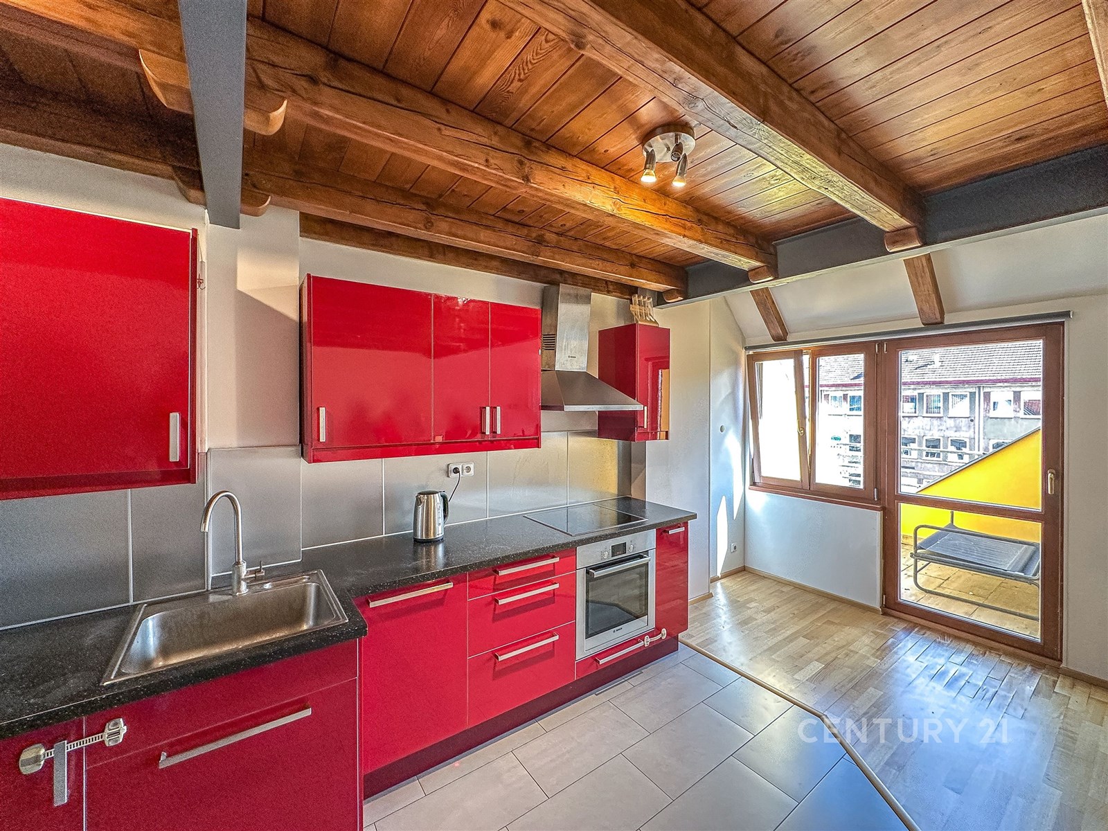 Moderní kuchyň s červenými lesklými skříňkami, dřevěným stropem a parketovou podlahou, velká okna poskytují dostatek světla