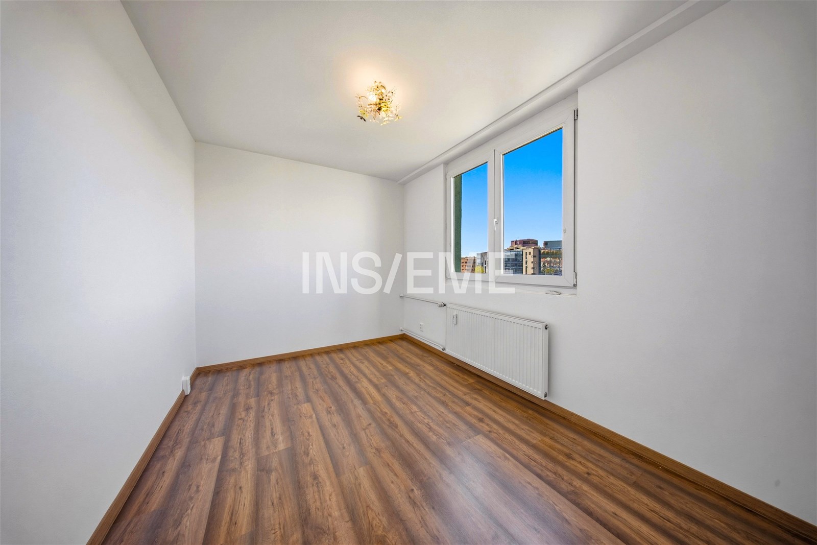 Prázdná ložnice s parketovou podlahou, velkým oknem a moderním osvětlením, nabízí světlý prostor ideální pro osobní přizpůsobení.