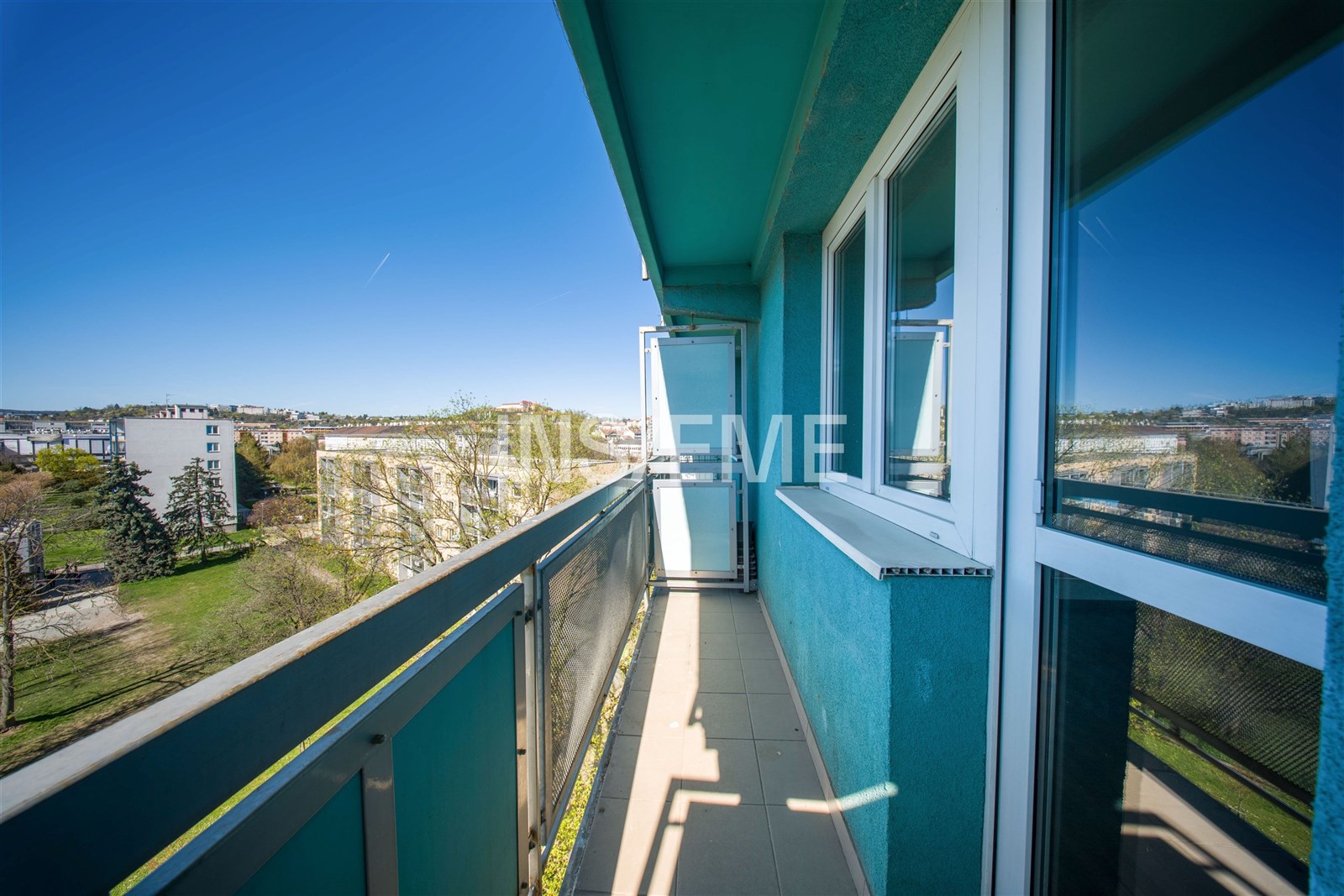 Úzký balkon s dlažbou a výhledem na zeleň a město, moderní zábradlí, přístup z interiéru velkými okny, světlé prostředí