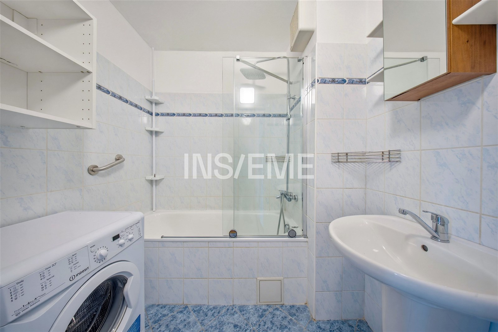 Koupelna s keramickou dlažbou, moderní sprchový kout s prosklenou zástěnou, pračka a vestavěné police pro úložný prostor.