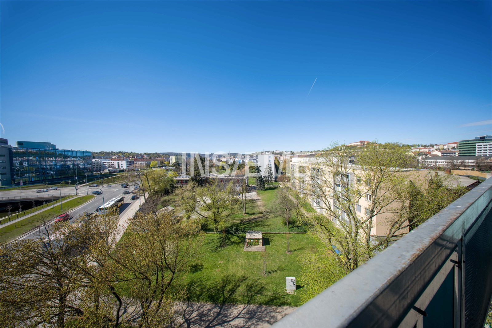 Výhled z balkonu na zelený park, okolí s moderními budovami a jasnou oblohou, ideální lokalita pro klidné bydlení ve městě.