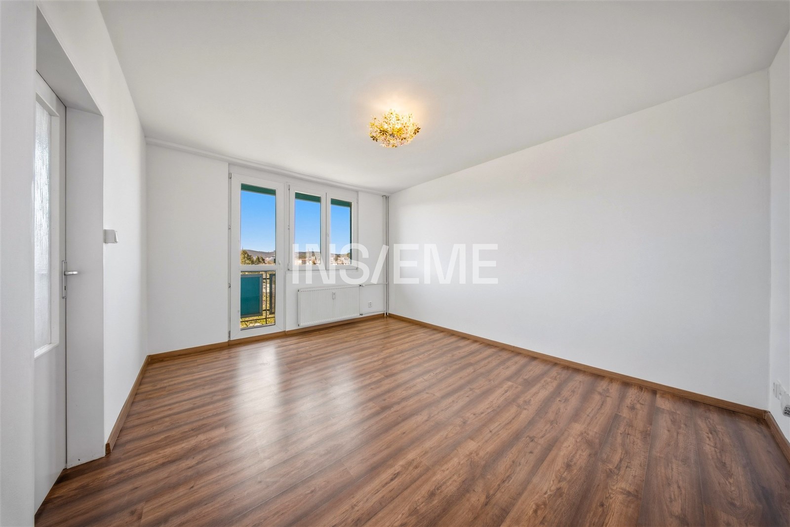 Světlý obývací pokoj s parketovou podlahou, velká okna a moderní lustr, v rekonstruovaném bytě.