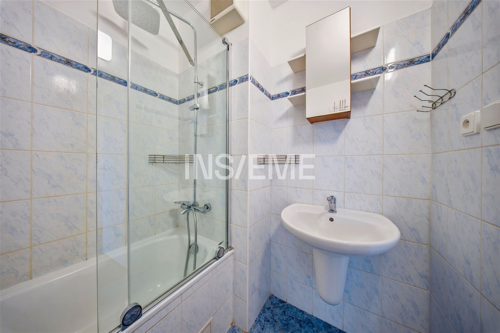 Koupelna s obklady, moderní sanitou, a prosklenou sprchovou zástěnou. Světlá modro-bílá dlažba doplňuje čistý a svěží vzhled.
