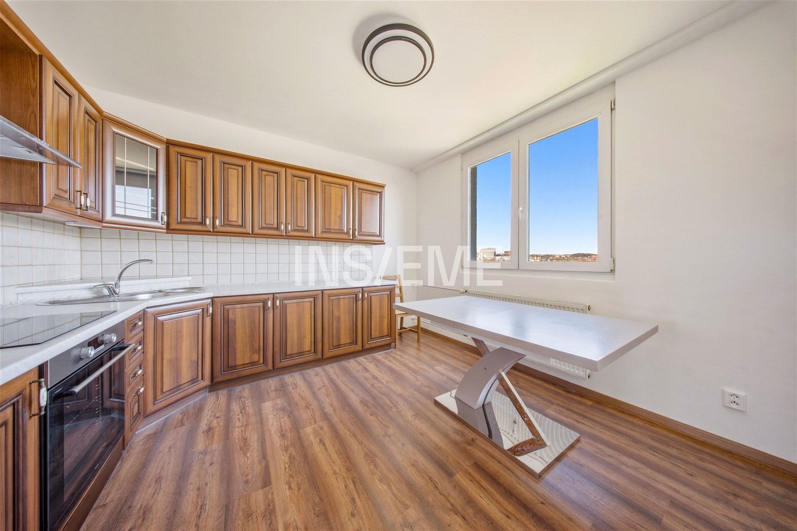 Moderní kuchyň s dřevěnými skříňkami, parketovou podlahou a velkými okny nabízí světlý a prostorný interiér vhodný pro rodinné vaření.